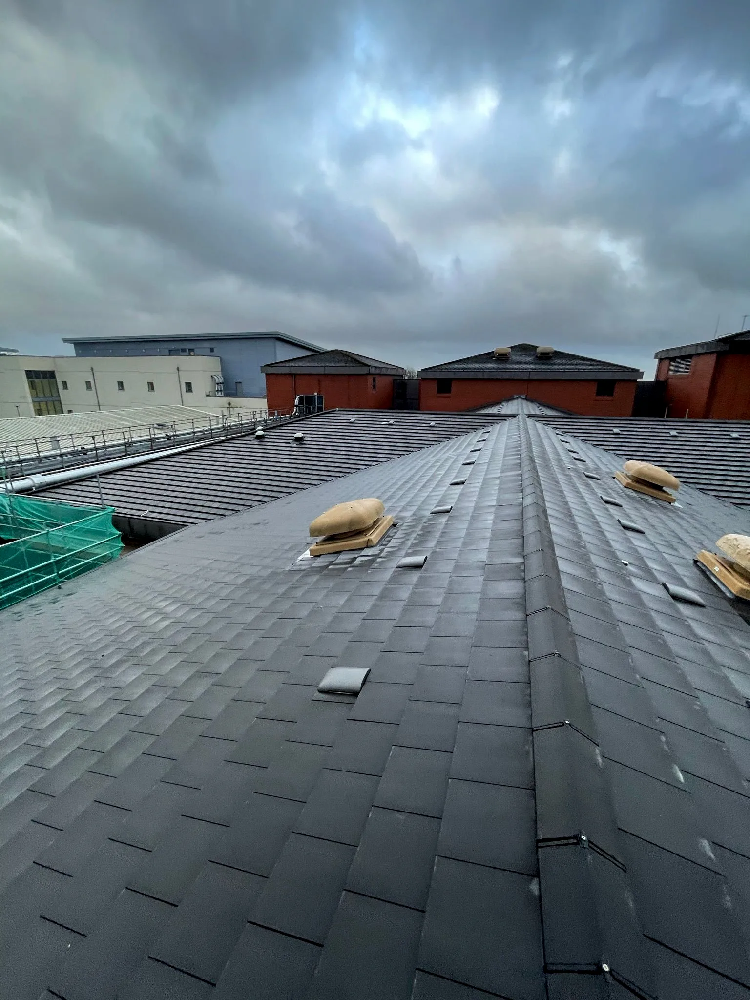 Newly installed dark slate-grey roof with ventilation hoods overlooking industrial buildings under stormy sky