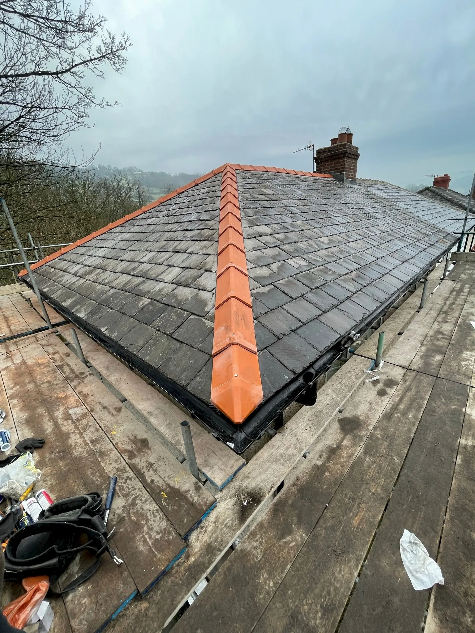 Newly installed dark grey slate roof tiles with bright orange ridge tiles and chimney, surrounded by scaffolding
