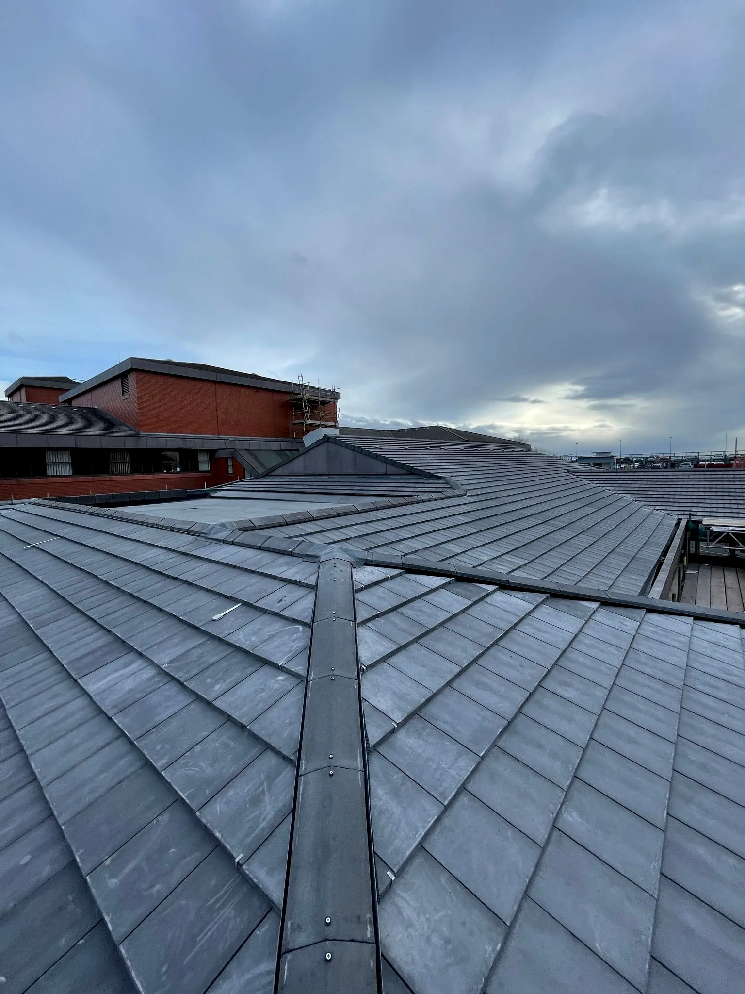 Large slate roof with central gutter and red brick building against cloudy sky